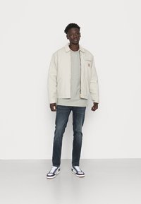 Light beige jacket with a zip, gray t-shirt underneath, dark blue jeans, and white sneakers with navy accents. Flat, smooth texture overall.