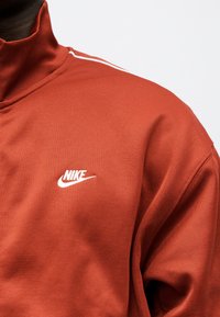 Κοντινό πλάνο ενός ατόμου που φοράει φούτερ Nike σε σκουρόχρωμη πορτοκαλί απόχρωση με λευκή επένδυση στον ώμο και λευκό λογότυπο Nike στο στήθος.