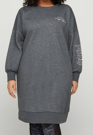 Person wearing a long, gray sweatshirt dress with text on the chest and right sleeve, paired with black and white patterned leggings.