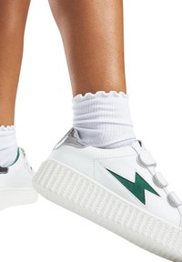 Sneaker blanche avec un design d'éclair vert, semelle texturée blanche, accents argentés, et trois sangles Velcro. Associée à des chaussettes blanches à côtes.