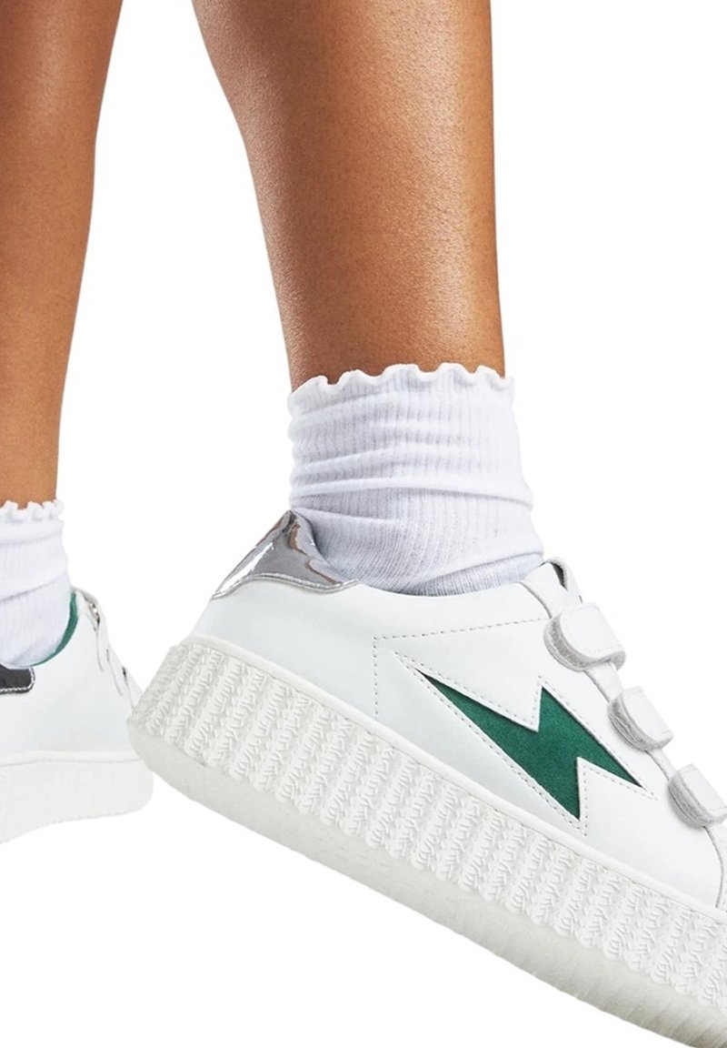 Sneaker blanche avec un design d'éclair vert, semelle texturée blanche, accents argentés, et trois sangles Velcro. Associée à des chaussettes blanches à côtes.