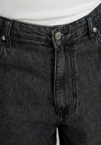 Jean en denim noir avec coutures apparentes et braguette à boutons, porté avec un t-shirt blanc côtelé rentré dans le pantalon.
