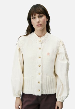 Vrouw draagt een crèmekleurig gebreid vest met knopen over een wit blouse met lange, opgeblazen mouwen, staand tegen een effen achtergrond.