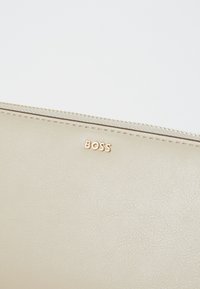 Beige kožená taška s texturovaným povrchem, na kterém se nachází zlatý logo "BOSS" a zlatý zipový uzávěr podél horního okraje.