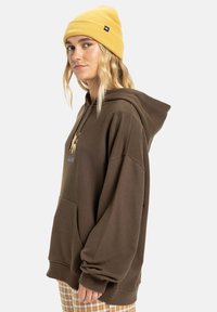 Brauner übergroßer Hoodie mit einer Fronttasche, verstellbarem Kordelzug und weißen Logo-Details. Kombiniert mit einer gelben gerippten Mütze.