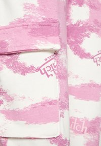 Tecido branco com padrão de pinceladas rosas, apresentando um bolso com aba com a palavra "Switch" impressa em rosa. Material suave e texturizado.