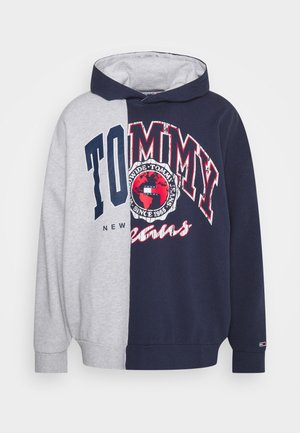 Väriblokillinen huppari, jossa on harmaat ja laivastonsiniset osiot, ja etupuolella iso "TOMMY"-logo. Yhdistetty vaaleansinisiin löysiin farkkuihin ja lenkkareihin.