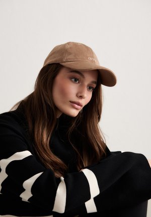 Jonge vrouw met lang bruin haar, een beige pet en een zwarte trui met witte patronen, die naar beneden kijkt tegen een effen achtergrond.