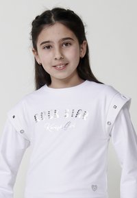 Fehér, hosszú ujjú ing, amelyen fényes ezüst szegecsekből van a "Punk Star" felirat. Fémes díszítésekkel rendelkezik a vállakon és egy kis szív logóval az alján.