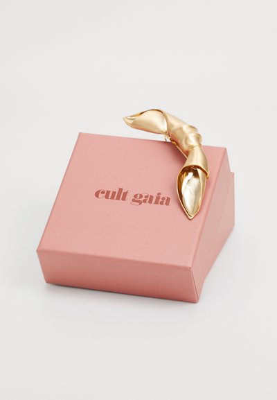 Gouden metalen haarclip met een gedraaide design, rustend op een roze vierkante doos met "cult gaia" in donkerroze geperst.