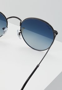 Ochelari de soare circulare cu ramă din metal negru, lentile gradient albastre, suporturi pentru nas transparente și brațe subțiri negre.
