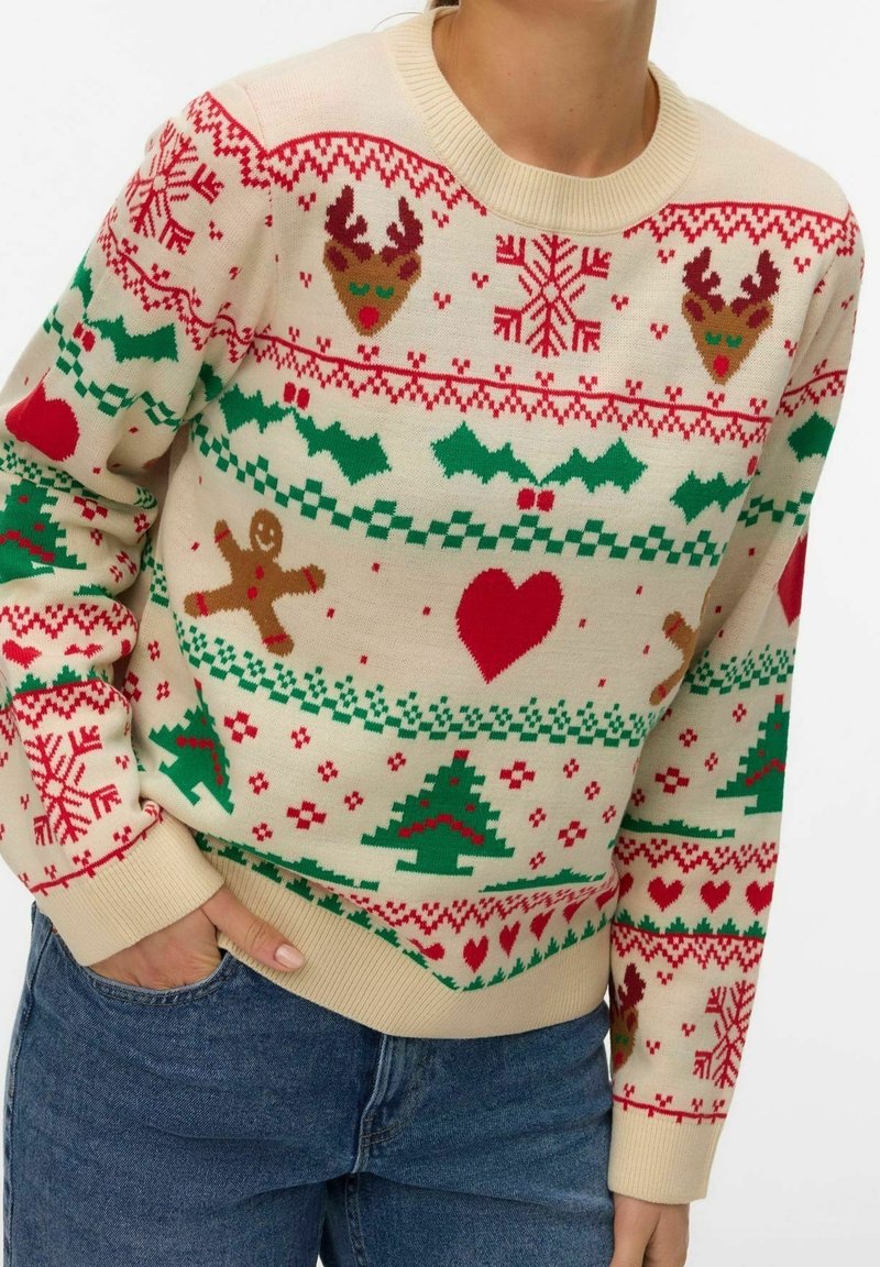 Personne portant un pull festif crème avec des motifs rouges et verts de vacances, comprenant des rennes, des flocons de neige, du houx, des cœurs, des bonshommes en pain d'épice et des sapins de Noël.
