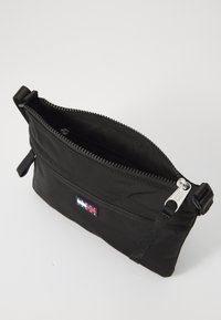 Svart nylon pouch med glidelås på toppen, som har en liten logo-patch og en avtakbar stropp for enkel bæring. Glatt tekstur og rektangulær form.