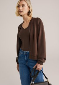 Barna cipzáros bomber dzseki bordázott gallérral, barna V-nyakú felsővel és világoskék, magas derekú farmerrel párosítva. Fekete kézitáskát tart.