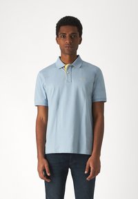 Polo shirt azzurro con placket a due bottoni, colletto giallo sul rovescio e delicata ricamata del logo. Indossato con jeans di denim scuro.