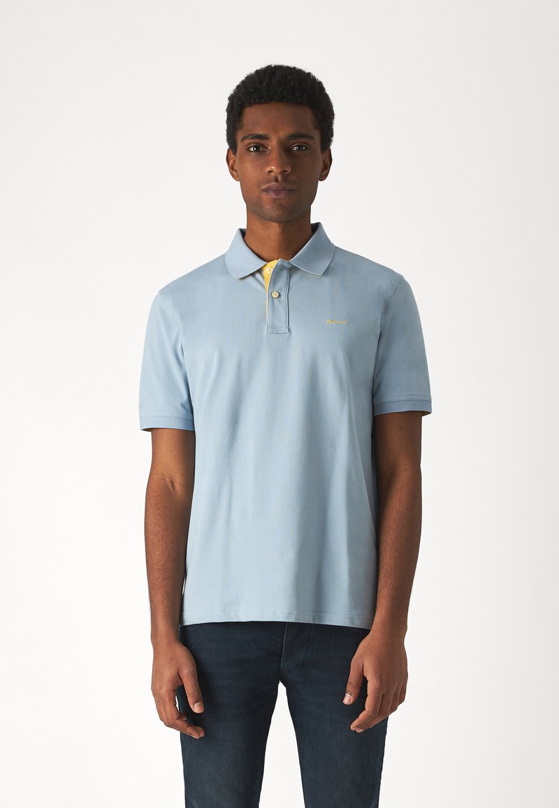 Polo shirt azzurro con placket a due bottoni, colletto giallo sul rovescio e delicata ricamata del logo. Indossato con jeans di denim scuro.