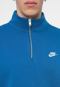 Μπλε φούτερ με φερμουάρ, φτιαγμένο από υφασμάτινη υφή, με ψηλό γιακά και λευκό λογότυπο Nike στο στήθος, καθώς και μεταλλική λεπτομέρεια στο φερμουάρ.