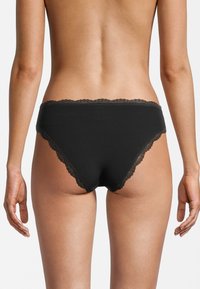 Donna vista di spalle che indossa biancheria intima nera in pizzo a coste, in piedi contro uno sfondo bianco semplice.