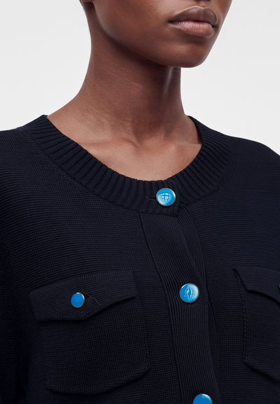 Gros plan d'une personne portant un cardigan en maille noire avec deux poches avant et des boutons décoratifs bleus présentant des motifs abstraits blancs.