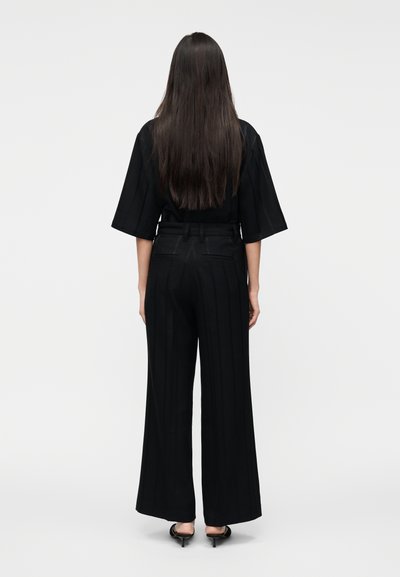 Juodos plačios kelnės aukštu liemeniu ir vertikaliomis juostomis, derinamos su atitinkama juoda palaidine. Audinys atrodo lygus su minimaliu blizgesiu.