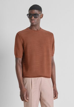 Uomo che indossa occhiali da sole neri oversize, maglione a maniche corte color ruggine e pantaloni beige chiaro su sfondo neutro.