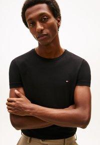 Črna kratka majica s kratkimi rokavi, okroglo izrezana, z majhnim večbarvnim logotipom na levi strani prsi. Gladek bombažni material.