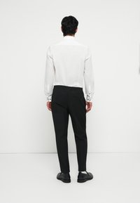Witte blouse met lange mouwen en contrasterende zwarte knopen, gecombineerd met zwarte broek in een slim fit met elastische tailleband. Zwarte schoenen.