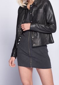 Zwarte leren jas met een drukknoopsluiting en borstzak, gecombineerd met een grijze, slim-fit jurk met een rits aan de voorkant.