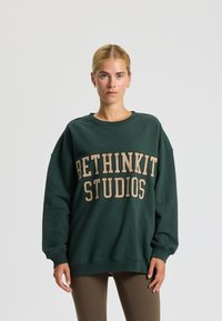Felpa verde con vestibilità ampia, colletto a girocollo a coste e stampa "BETHINKIT STUDIOS" in beige. Tessuto morbido e maniche lunghe.