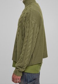 Oliwkowy sweter robiony na drutach z wysokim kołnierzem i wzorem w warkocze, z prążkowanymi mankietami i luźnym krojem. Nosi się go na dopasowanej zielonej koszuli.