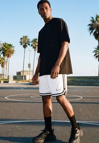 Mlad moški stoji na zunanjih košarkarskih igriščih, oblečen v črno majico, bele kratke hlače, črne nogavice in črne superge, v ozadju so palmova drevesa.