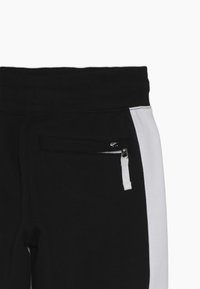 Svart träningsoverall med en vit sidopanel, elastisk midja och en ficka med dragkedja och detaljer i metall. Tyget ser mjukt och avslappnat ut.