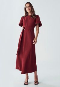 Robe midi rouge avec de courtes manches bouffantes, une ceinture sur le côté avec une boucle circulaire, et une texture lisse, associée à des sandales noires à lanières.