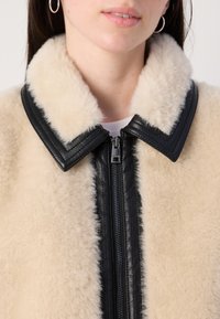 Shearling jack met een crèmekleur en een zwarte leren kraag en rits. De stof heeft een zachte, textuurrijke uitstraling.
