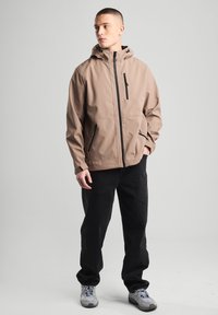 Light brown insulated jacket with a hood, front zipper, and two side pockets, paired with black pants and gray sneakers.