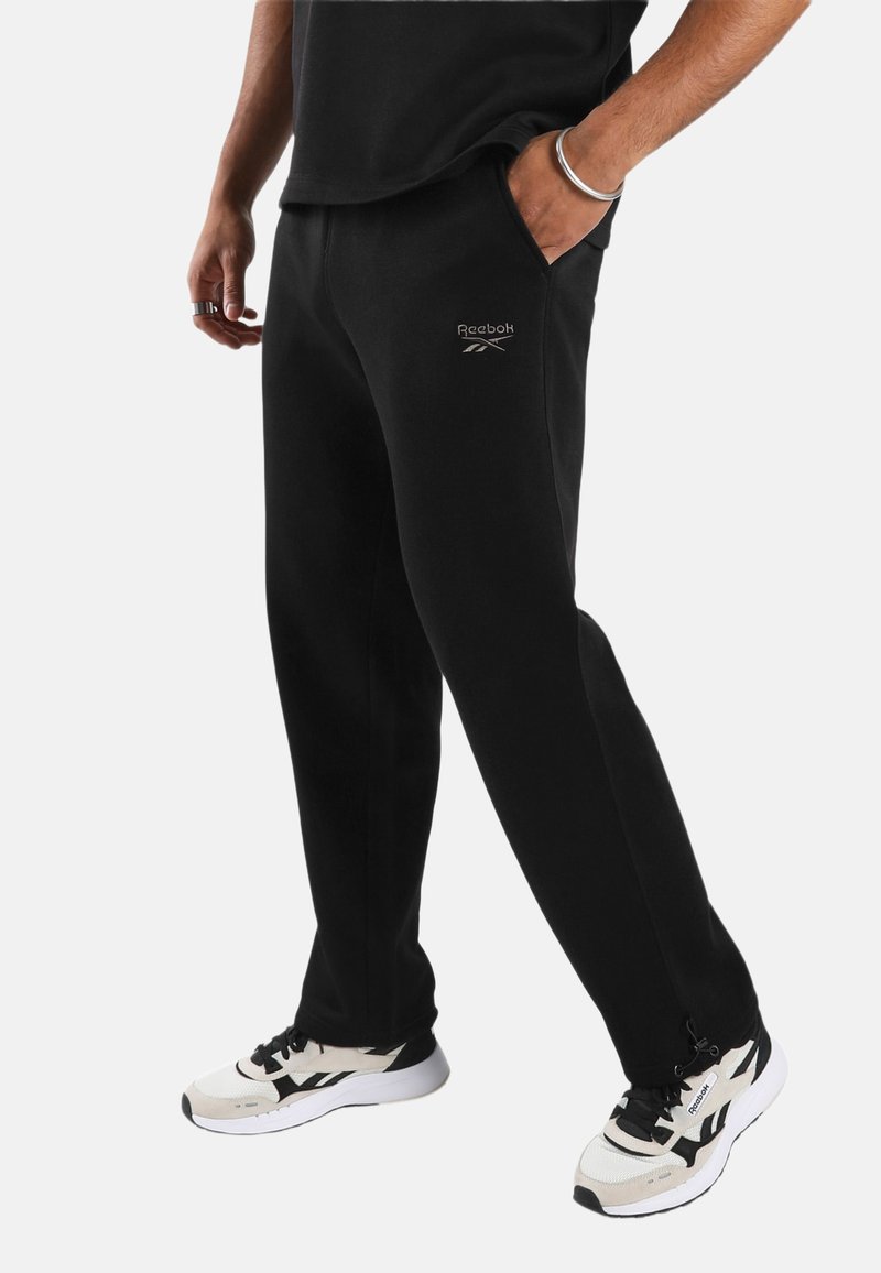 Person trägt schwarze Reebok-Sweatpants und weiße-schwarze Reebok-Sneaker und steht vor einem schlichten, hellen Hintergrund.