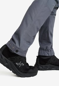 Svarta sportskor med grå logotyp, använda med grå byxor med tryckknappar vid anklarna mot en vit bakgrund.