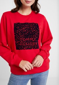 Rode sweatshirt met een marineblauw vierkant grafisch ontwerp met een bloemenpatroon en de tekst "TOMMY HILFIGER" in vette letters. Ribgebreide manchetten en onderrand.