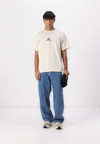 Beige cotton t-shirt with a blue graphic logo, paired with loose blue denim jeans and beige sneakers. Accessories include a black bag.