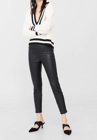 Kobieta stoi z założonymi rękami, ubrana w białą sweter V-neck w czarne paski, czarne skórzane spodnie oraz czarne czółenka na niskim obcasie.