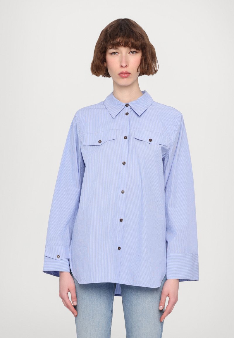 Camicia a righe blu con colletto, maniche lunghe, due tasche sul petto e bottoni di colore marrone. Realizzata in tessuto leggero.