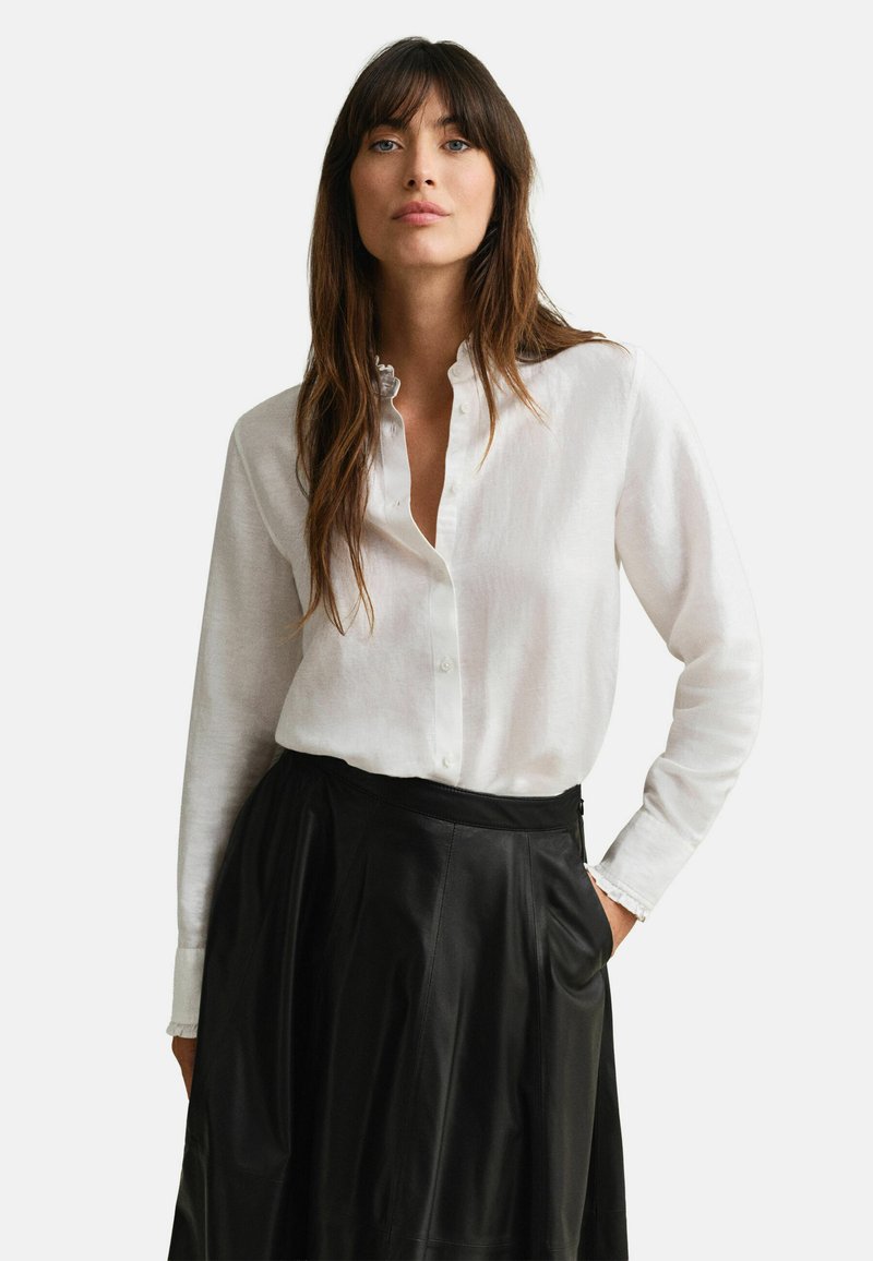 Femme aux longs cheveux bruns portant un chemisier blanc boutonné et une jupe en cuir noire, une main dans la poche, sur un fond clair uni.