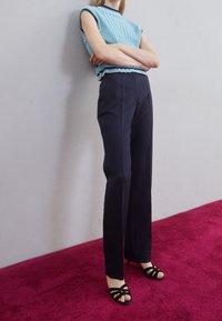 Jasnoniebieski top z żebrowanej dzianiny z falbaną na dole, sparowany z dopasowanymi granatowymi spodniami. Modelka stoi na fuksjowym dywanie w czarnych sandałach na obcasie. 