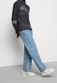 Tumma laivastonsininen neulottu neule, jossa on kuvioituja kuvioita, vaaleansiniset löysät farkut ja harmaanvalkoiset tennarit, joissa on raidalliset yksityiskohdat.