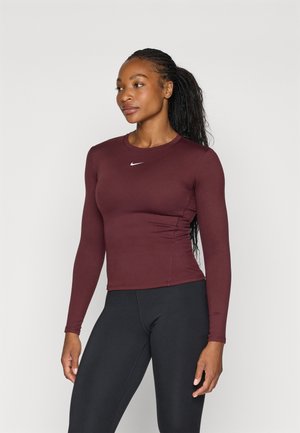 Burgundi színű, testhezálló, hosszú ujjú sportfelső, kerek nyakkivágással, középen Nike logóval és strukturált anyagból készült. Fekete leggingsszel párosítva.