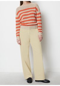 Osoba ubrana w beżowe spodnie o szerokich nogawkach, czarne mokasyny i beżowy sweter w poziome pomarańczowe paski, stojąca przy białej ścianie.