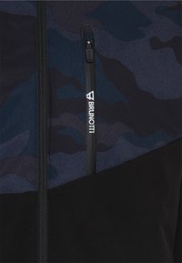 Primo piano di una giacca blu scuro e nera con motivo camo e una tasca verticale con zip che mostra il logo Brunohti.