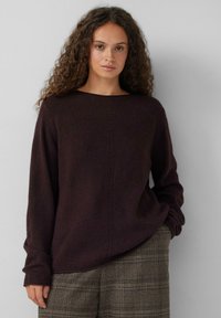 Vrouw met krullend haar in een donkerbruine trui en een geruite broek, staande tegen een effen lichtgrijze achtergrond.