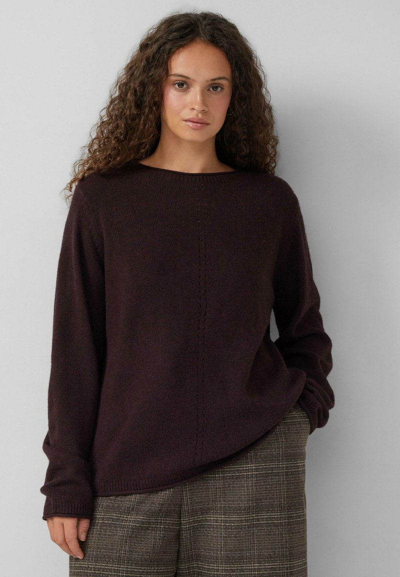 Vrouw met krullend haar in een donkerbruine trui en een geruite broek, staande tegen een effen lichtgrijze achtergrond.