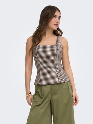 Jeune femme portant un haut sans manches à carreaux marron et blanc et un pantalon ample vert olive, regardant sur le côté avec de longs cheveux bruns.